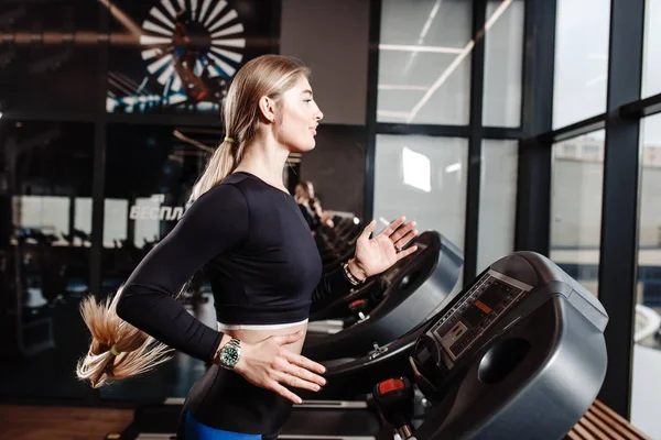 The athletic girl dressed in a sportswear is running on the treadmill in front of the windows in the modern gym