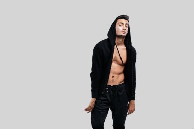 Şık genç dansçı bir kapüşonlu ve siyah pantolon ile çıplak bir gövde üzerinde bir sweatshirt giymiş beyaz bir arka plan üzerinde duruyor
