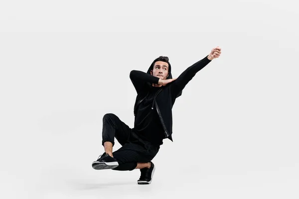 Dançarina elegante de cabelos escuros vestida com uma roupa preta faz movimentos estilizados de dança de rua. Ele está sentado em uma perna e colocar a mão para cima — Fotografia de Stock
