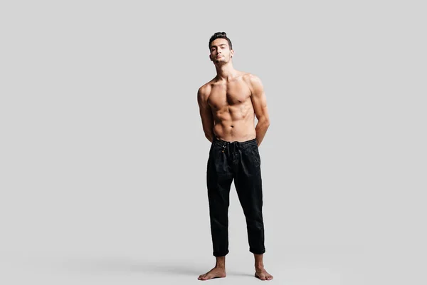 Dark-haired handsome young dancer with bare torso wearing a black sports pants is standing on a white background — Stock Photo, Image
