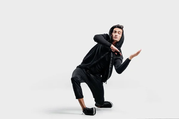 Dançarino elegante de cabelos escuros vestindo roupas pretas e um capuz faz movimentos estilizados de dança de rua . — Fotografia de Stock