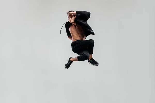 Handsome young dancer of street dancing dressed in black pants and a sweatshirt on a naked torso jumps on a white background