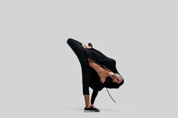 Young handsome young dancer dressed in black pants, a sweatshirt on a naked torso dances breakdance holding one hand and one foot on the floor — Stock Photo, Image