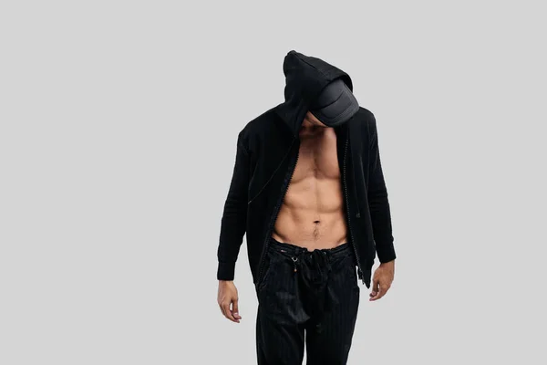 Bonito jovem dançarino vestido com uma camisola em um tronco nu com um capuz na tampa e calças pretas fica em um fundo branco — Fotografia de Stock