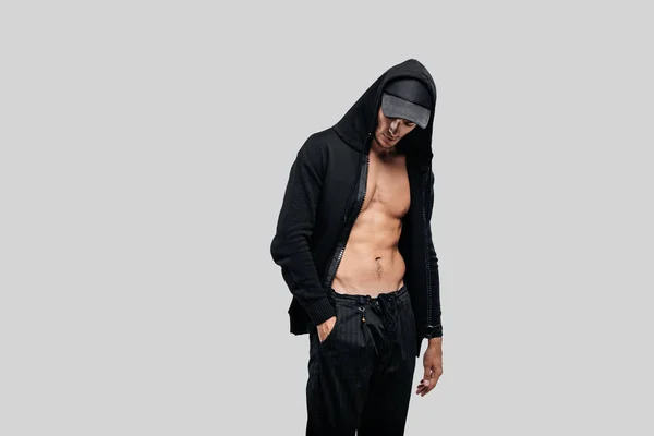 Bonito jovem dançarino vestido com uma camisola em um tronco nu com um capuz no boné e calças pretas fica com a mão no bolso — Fotografia de Stock