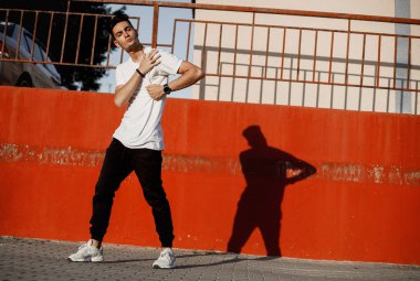 Genç adam kot pantolon ve beyaz t-shirt giymiş güneşli bir gün boyalı beton duvara karşı sokakta modern dans dans ediyor