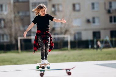 Komik çocuk şık rahat giysiler giymiş güneşli sıcak bir günde evin yanında bir platformda kaykay sürmek