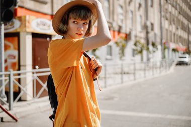 Sarı bir t-shirt ve hasır şapka genç kız bir yaz güneşli bir günde bir şehir caddesi boyunca bir sırt çantası ile yürüyor