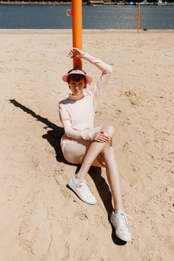 Hafif pembe elbise ve pembe vizör şık kız bir yaz güneşli bir günde bir sütun üzerinde eğilerek kumlu bir plajda oturur