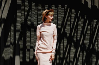 Yansıyan gölgeler ile güneş ışığında koyu bir metal çit yanında duran hafif pembe elbise ve hasır şapka şık kız