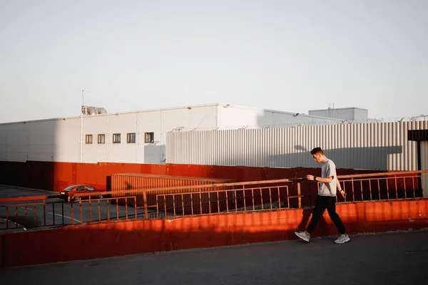 Jovem cara elegante vestido de jeans e t-shirt branca está andando ao lado do edifício urbano com estacionamento no pôr do sol — Fotografia de Stock