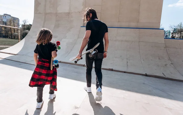 Vader en zijn kleine zoon gekleed in de stijlvolle casual kleren lopen met de Skateboards in hun handen in een skatepark op de zonnige dag — Stockfoto