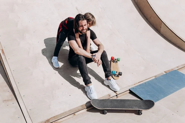 Gelukkig jonge vader en zijn zoon gekleed in de stijlvolle casual kleding zitten in een omhelzing samen op de glijbaan naast de Skateboards in een skatepark op de zonnige warme dag — Stockfoto
