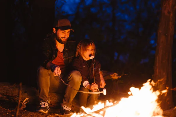 Happy father and his little son sitting on the logs in the forest in front of a fire and eating marshmallows from the sprigs