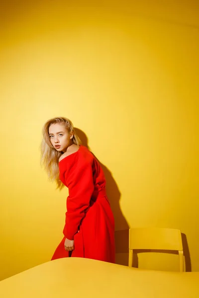 V elegantní červené kabátě stojí na žlutém stole na pozadí žlutých stěn módní blogovací dívka. — Stock fotografie