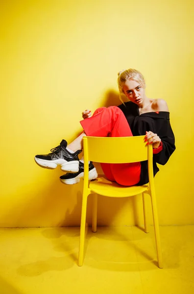 Mladá módní dívka blogger oblečená v červených kalhotách a černém Jacku, která sedí na stoličce v místnosti se žlutými stěnami a nábytkem — Stock fotografie