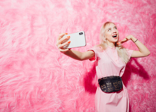 Bright young girl blogger dressed in fashion pink dress takes a selfie on her smartphone on the background of pink fur wall in the show room Stock Image