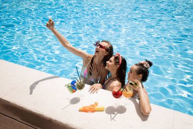 Mayolar içinde Ağaç şık genç kızlar rahatlatıcı ve güneşli bir yaz gününde açık havada yüzme havuzunda kokteyller ile salfie yapma