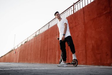 Kot giymiş genç adam ve t-shirt şehirde yaz gününde bir boyalı beton duvara karşı bir scooter ile duruyor.