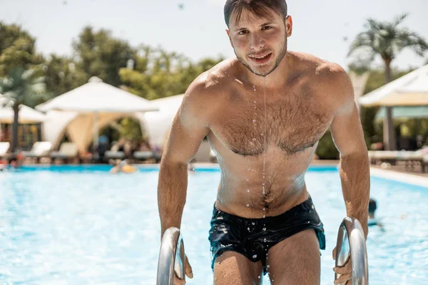 Jovem bonito emerge da piscina apoiada no corrimão das escadas na piscina ao ar livre em um dia ensolarado de verão — Fotografia de Stock