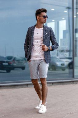 Şık adam rahat tarzı ve güneş gözlüğü bir yaz günü bir cam binanın arka planına karşı poz moda yazlık giysiler giymiş