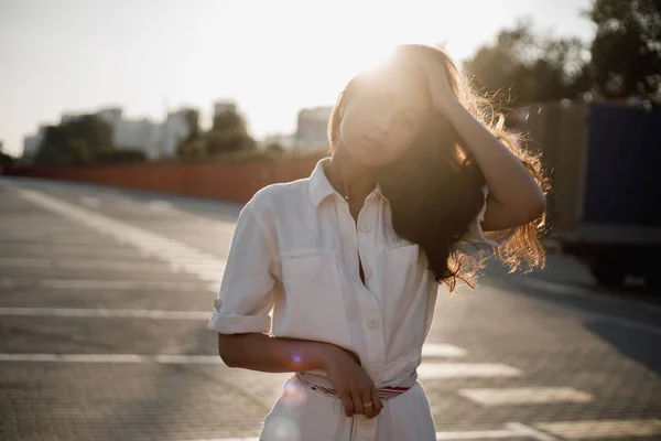 Giovane ragazza con lunghi capelli scuri vestita con camicia bianca è in piedi in strada sotto il sole luminoso — Foto Stock