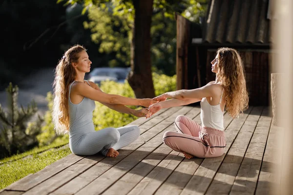 Twee jonge vrouwen zitten en houden handen in de lotuspositie op een houten podium in de tuin in de buurt van het huis in de zonnige ochtend — Stockfoto