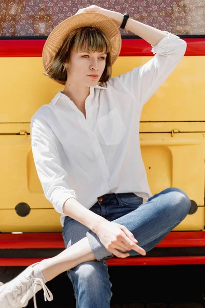 Стильна молода жінка в солом'яному капелюсі, одягнена в білу сорочку і джинси, сидить на бампері яскраво-червоного і жовтого мінівена — стокове фото