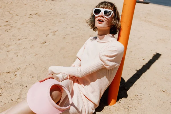 Menina elegante em óculos de sol brancos, um vestido suavemente rosa e viseira rosa senta-se em uma praia de areia apoiada em um pilar em um dia ensolarado de verão — Fotografia de Stock