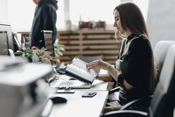 Tineri bruneta fata imbracata in haine de birou stil este așezat la birou și păstrează notebook-ul în mâinile ei un birou modern lumină — Fotografie, imagine de stoc