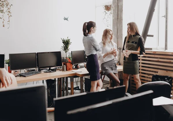 Three beautiful young women dressed in office style clothes stand together and talks in the modern office