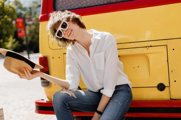 Élégante jeune femme aux lunettes de soleil blanches vêtue d'une chemise blanche et d'un jean tenant un chapeau de paille à la main s'assoit sur le pare-chocs d'un minifourgon rouge et jaune vif — Photo