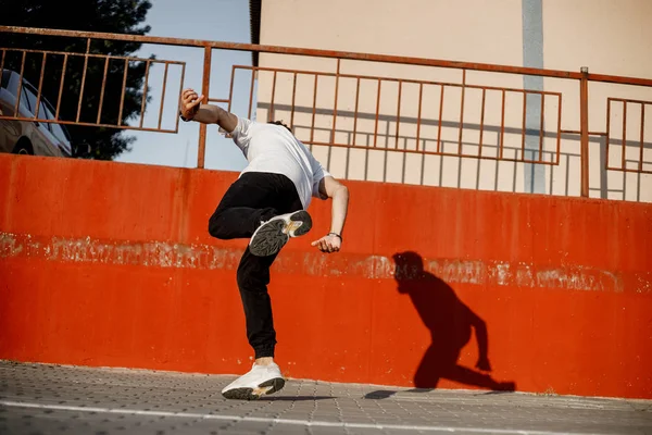 Jonge kerel gekleed in jeans en wit t-shirt is dansen moderne dans in de straat tegen een geschilderde betonnen muur in de zonnige dag — Stockfoto