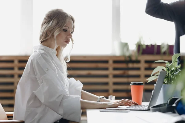 Ung, vakker kvinne i hvit skjorte jobber på laptopen ved skrivebordet i et moderne, åpent romkontor. – stockfoto