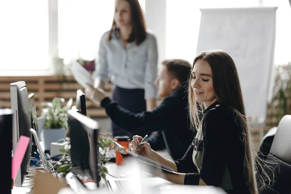 Unge brunettpike kledd i kontorklær sitter ved skrivebordet med kollegene og har notisboken i hendene i et lett, moderne kontor. – stockfoto