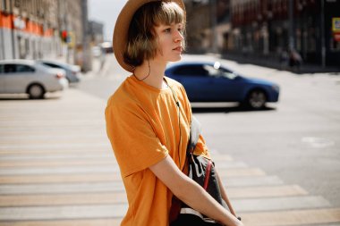 Sarı bir t-shirt ve hasır şapka genç kız bir yaz güneşli bir günde bir şehir caddesi boyunca bir sırt çantası ile yürüyor