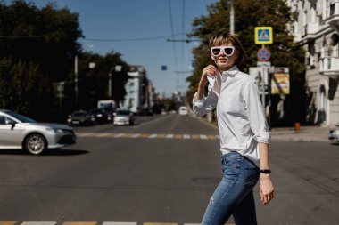 Beyaz gömlek ve kot pantolon giymiş beyaz güneş gözlüğü şık genç kadın bir yaz güneşli bir günde bir şehirde geçiyor