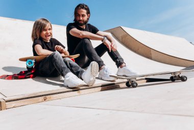 Genç baba ve oğlu şık rahat giysiler giymiş güneşli sıcak bir günde bir kaykay parkta kaykaylar yanında slayt üzerinde birlikte oturuyor