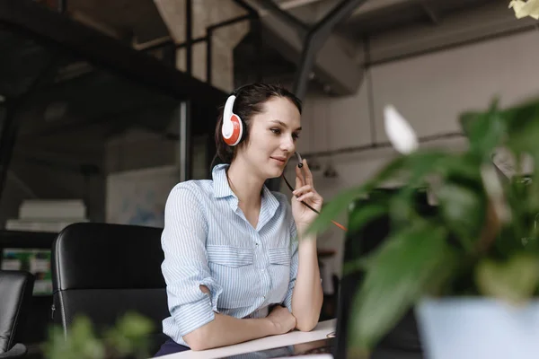 Ung brunett kvinna klädd i kontors stil kläder pratar med kunder genom headsetet sitter vid Skriv bordet med laptop i det moderna kontoret — Stockfoto