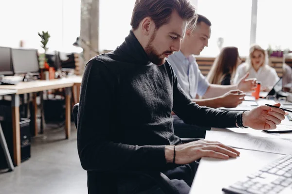 Tipul îmbrăcat în haine de birou casual lucrează cu documente care stau la birou cu computerul lângă colegii din biroul modern de lumină — Fotografie, imagine de stoc