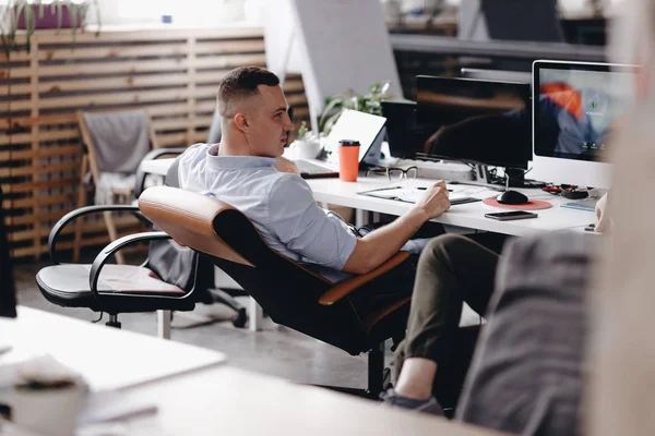 The guy dressed in office style clothes is sitting in an office chair at desk with laptopo in the modern office