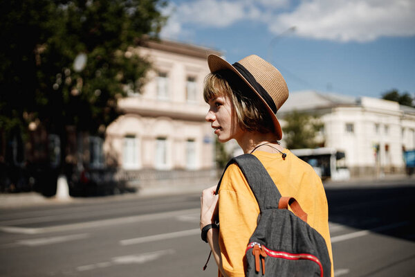 Young girl with headphones in a yellow t-shirt and a straw hat crossing the road with a backpack in a city street on a summer sunny day