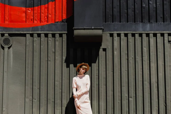 Menina elegante em um vestido suavemente rosa e chapéu de palha em pé ao lado de uma cerca de metal escuro em um dia ensolarado — Fotografia de Stock