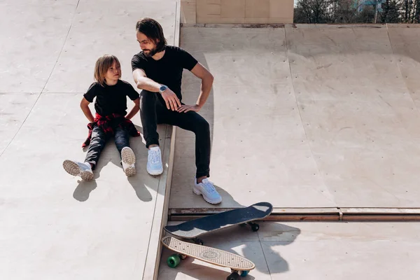 Happy Father en zijn zoon gekleed in de casual kleding zitten en lachen op de glijbaan in een skatepark naast de Skateboards op de zonnige dag — Stockfoto