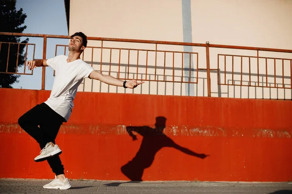 Jovem vestido de jeans e camiseta branca está dançando dança moderna na rua contra uma parede de concreto pintada no dia ensolarado — Fotografia de Stock