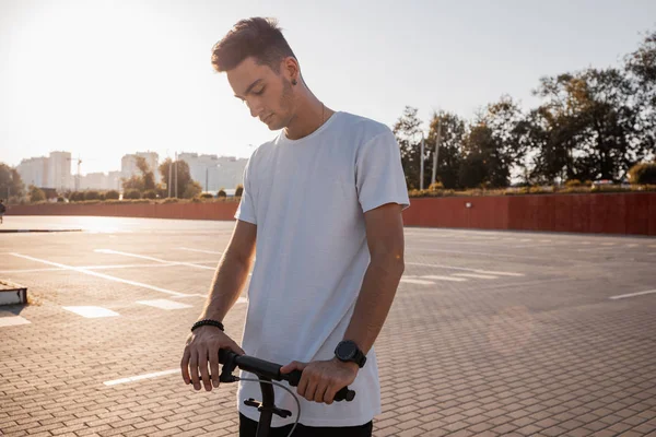 Jovem vestido de jeans e t-shirt está montando uma scooter na praça para estacionamento no dia ensolarado — Fotografia de Stock