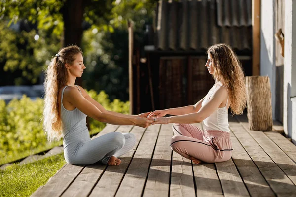 Due giovani donne si siedono e si tengono per mano nella posizione del loto su un podio di legno nel giardino vicino alla casa all'aria aperta — Foto Stock