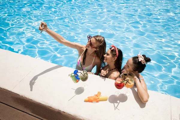 Mayolar içinde Ağaç şık genç kızlar rahatlatıcı ve güneşli bir yaz gününde açık havada yüzme havuzunda kokteyller ile salfie yapma