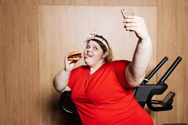 Fat and funny girl dressed in the sportswear and with a bandage on her head is making selfie standing with burger in her hand next to the treadmill