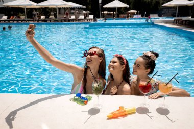 Mayolar içinde Ağaç şık genç kızlar rahatlatıcı ve güneşli bir yaz gününde açık havada yüzme havuzunda kokteyller ile salfie yapma